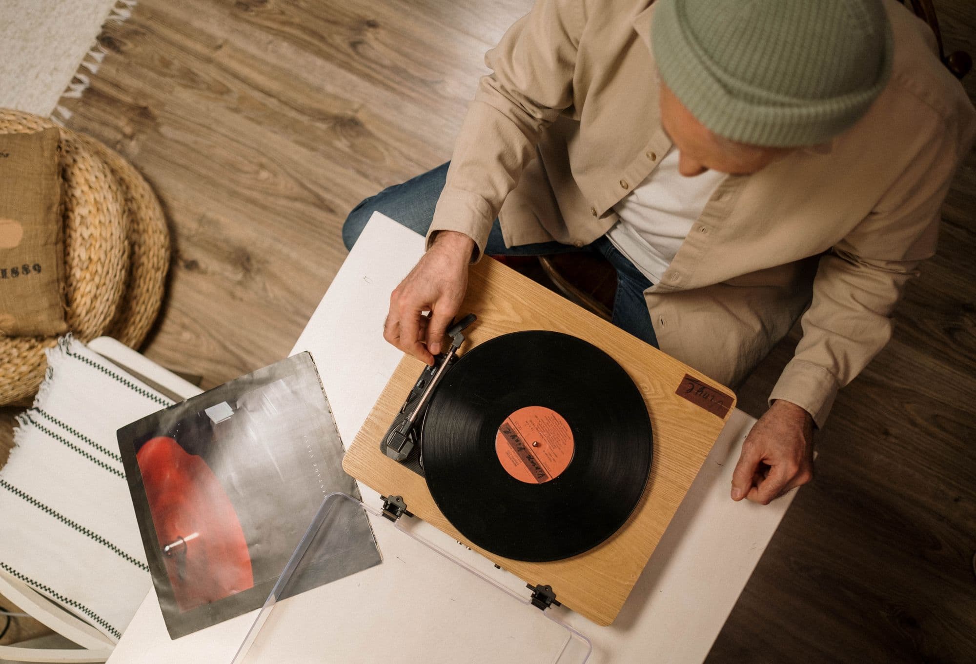 Overhead view of a person cueing a classic album on a wooden turntable, an album sleeve resting on the floor beside them.