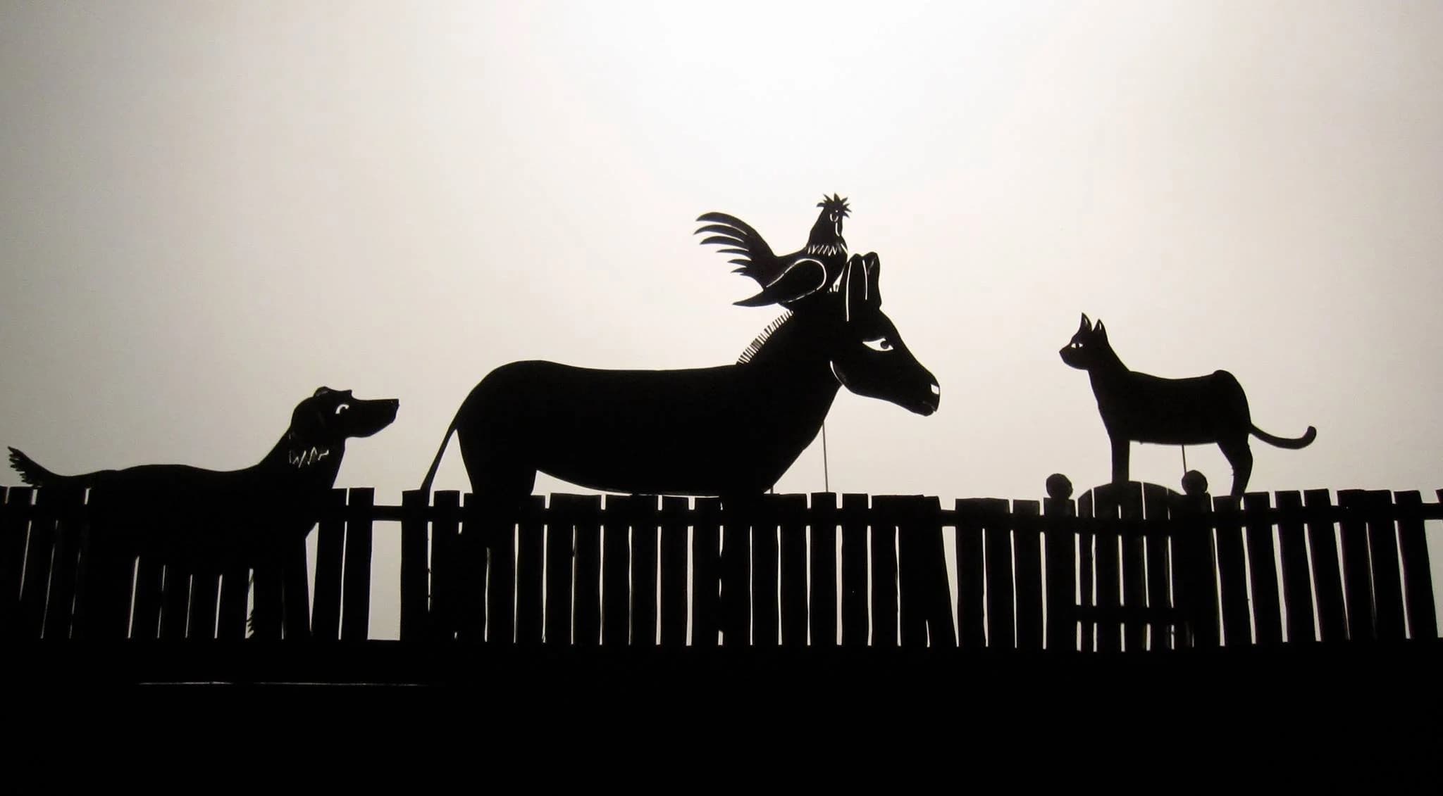 Shadow-puppet silhouettes of the Town Musicians of Bremen — a dog, a donkey with a rooster on its head, and a cat — atop a fence, by CactusHead Puppets.